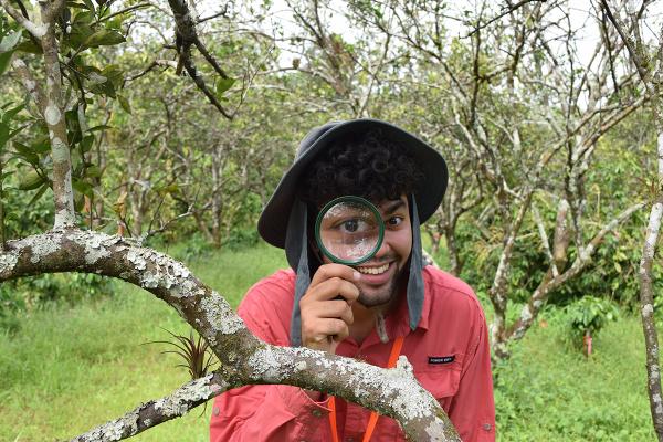 Jacob Longmeyer outside looking at the camera through a magnifying glass, smiling, near a tree where he was observing ants