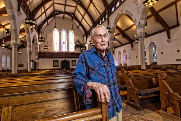 Jim Toy stands at a pew at St. Andrew's Episcopal Church, a light smile lingering on his face. There is a glint in his eyes, the sleeves of his blue plaid shirt rolled up.