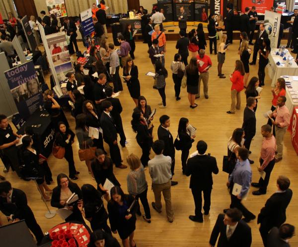 A room full of students who are dressed in business / business casual attire. The students are in lines to speak with various organizations as part of a career fair.