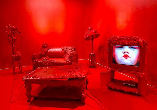 Furniture decorated with organic-looking additions is shown in a room. All visible surfaces are painted deep red with the exception of a television screen that shows a face wearing pale makeup and red lipstick.