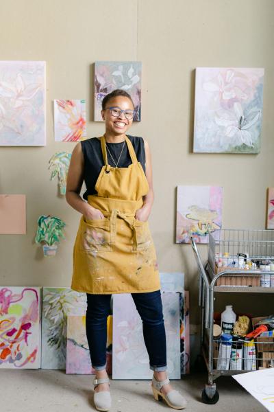Artist Avery Williamson, standing, wearing yellow apron surrounded by art canvases