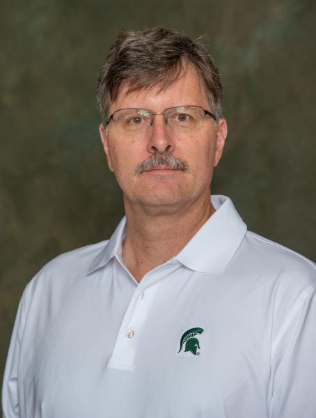 Jeff Freymueller, white man with glasses, brown hair and mustache wearing a white Michigan State polo