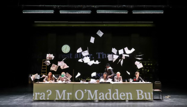 Theater company Elevator Repair Service on stage performing scenes from Ulysses. Five people sitting at a long table with papers flying all around. Text from the book can be seen displayed across the table and in the background.