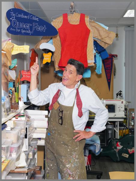 Phranc points to a sign saying "The Cardboard Cobbler: Queer feet are our specialty!"