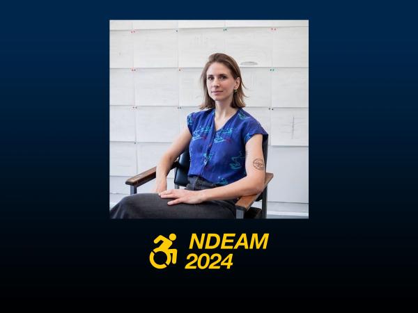 A white woman with chin length hair sits in a chair looking at the camera. She has a blue short sleeved shirt on and dark pants. Behind her are many drawings pinned to a wall. NDEAM 2024 logo.