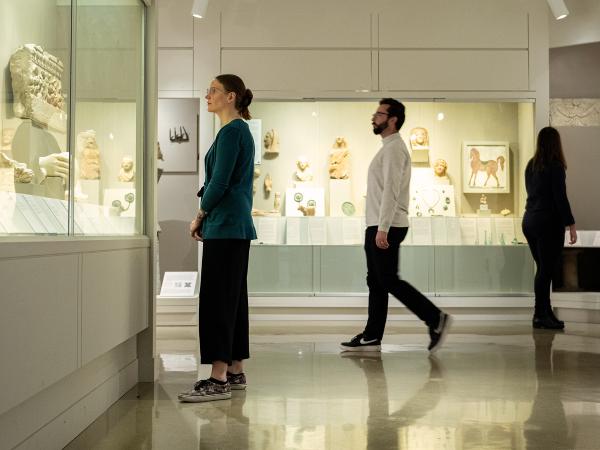A view of the Kelsey Museum’s Roman galleries. At the left, a woman looks at marble architectural fragments; in the center, a man passes by the cases; and at the right, a woman looks at objects related to Roman baths.