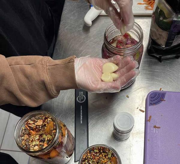 Two mason jars being filled with vegetables and spices to make pickles on a stainless steel counter top