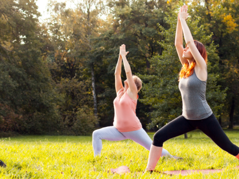 Two people doing a yoga pose outside with bent legs and their arms above their heads