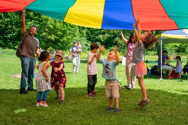 A group of young children play under a colorful parachute that is being held up by smiling adults in Nichols Arboretum