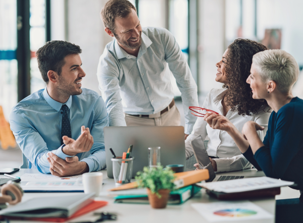 A group of business professionals, both men and women, smile and talk with each other at a table with a laptop, pens, and paper.