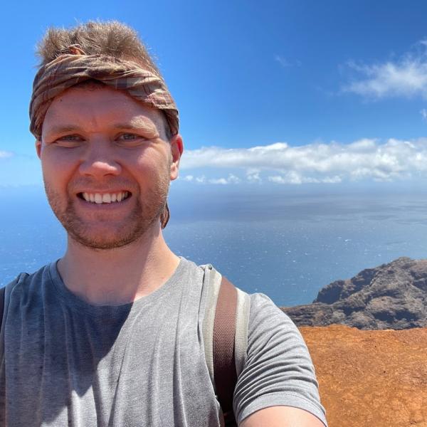 Photograph of Andrew Heard wearing a grey t-shirt with a brown bandana headband. Background: water with blue sky and clouds.