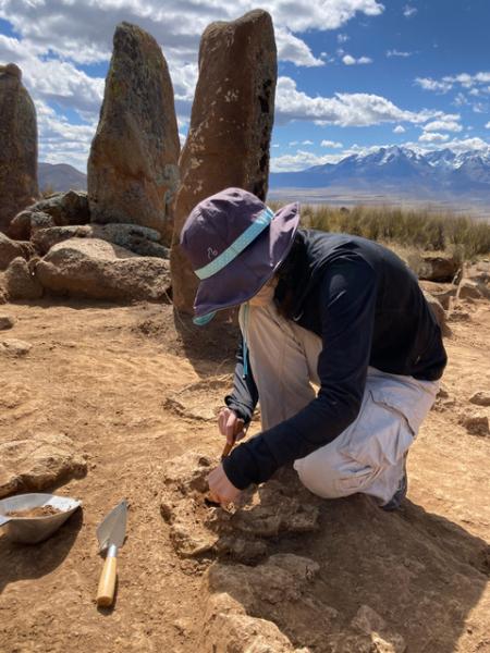 Man wearing black shirt, purple hat and tan pants crouched down on light brown ground using a tool to carefully dig
