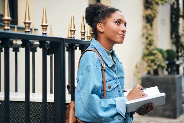 Side view of casually dressed African American student girl in denim jacket with backpack dreamily writing notes on city street