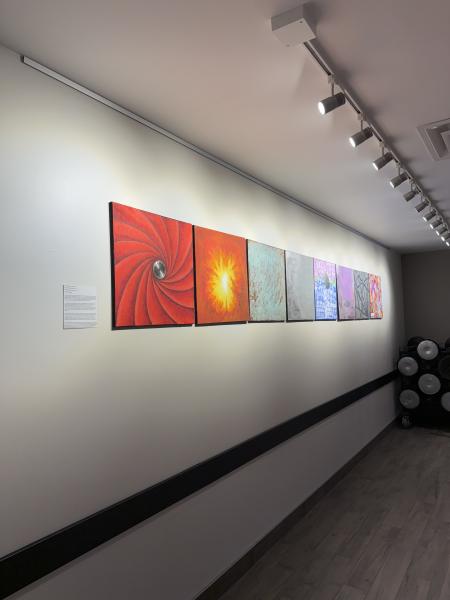 8 pieces of art on a white wall with ceiling lighting illuminating exhibit.