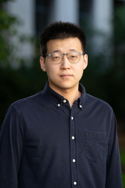Photograph of Yaolin Miao weraing a dark blue button up shirt with white buttons, and grey-framed glasses.