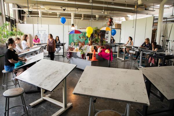 Students draw at white tables surrounding a colorful still life.