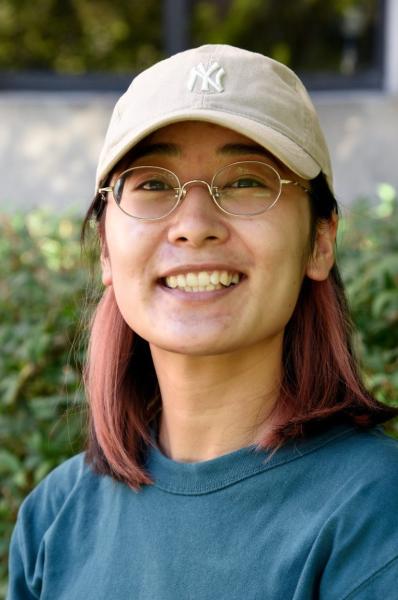 Photo of a girl with brown hair and glasses wearing a teal shirt and a tan New York hat smiles at the camera