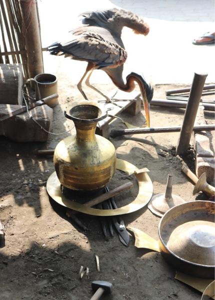 A purple heron stands among metalworking tools and vessels in a sunlit, earthen-floored workspace.