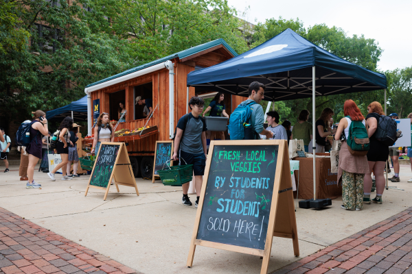 A mobile farm stand trailer with students gathered around, surrounded by tented booths. Signs in front read ‘Farm Stand’ and ‘Fresh, local veggies by students for students sold here!’ Students carry baskets of produce and chat with Farm Stand employees behind the tables, with trees and brick walkways in the background.