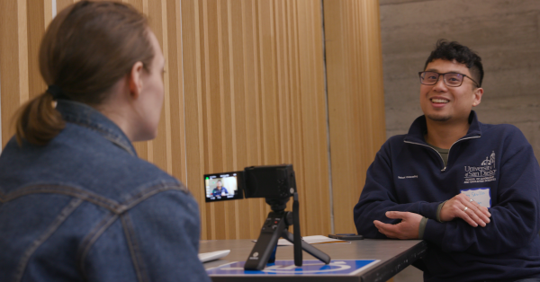 Two students sit across from each other, engaged in conversation, while one films the interview