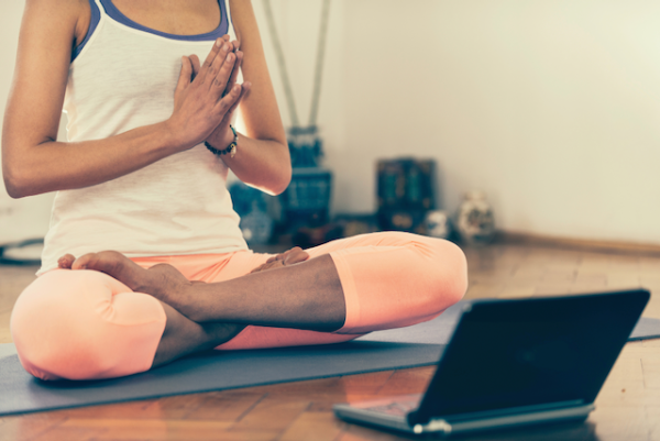 Woman sitting on the floor on a yoga mat, with her legs folded, hands together, in front of an open laptop.