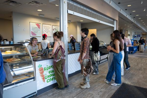 This image depicts a line of people waiting to order in front of a cafe counter. The counter reads "Rooting for Change Cafe."