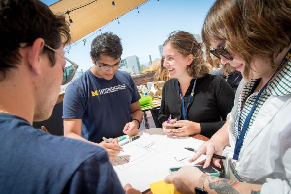 A group of students sit around a table, collaborating on a project. They are sitting outdoors in sunny weather and smiling at each other.