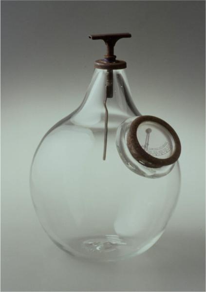 A clear, hand-blown glass sculpture or vessel by artist Marianetta Porter, featuring a rounded body, a dark metal "T" handle at the top neck, and a vintage-style dial or gauge integrated into a side opening.