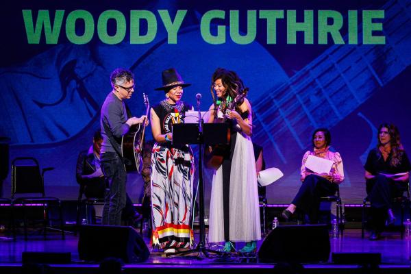 Three musicians perform with acoustic guitars on a purple-lit stage below large green text reading "WOODY GUTHRIE".