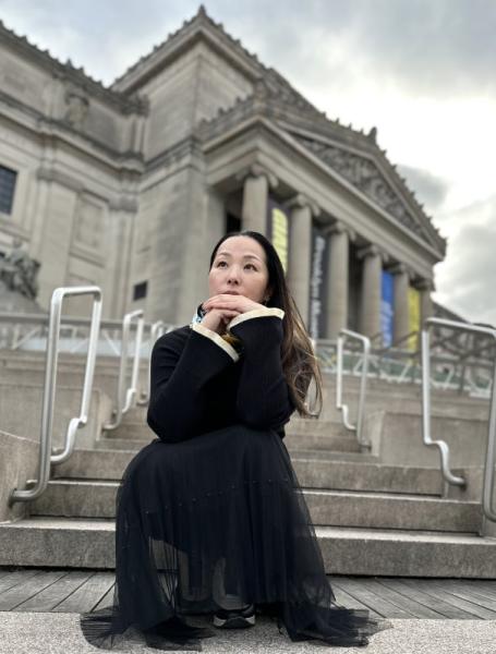 Kanako Uzawa sits outside of a large gray museum-like building, looking pensive up at the sky.