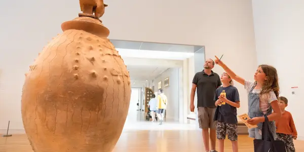 A large, textured clay pot stands in a museum gallery. A group of children and an adult look at the artifact, with one child pointing upwards and holding a brochure.