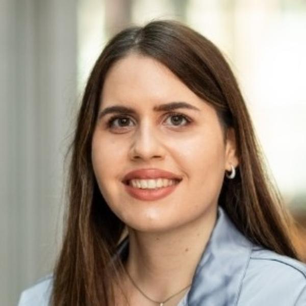 Headshot of a smiling woman with long straight dark hair, wearing small hoop earrings and a light blue top against a soft, out-of-focus background.