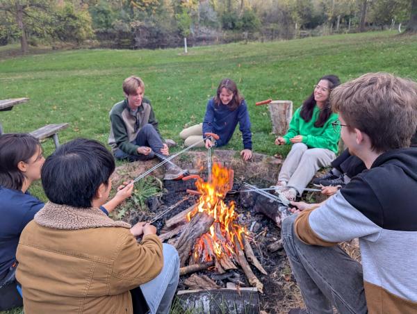 Students sit around a campfire pit roasting marshmallows in a green woody area