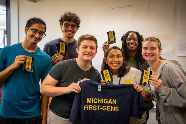 Six students standing in a line holding stickers that have a first-gen flag and two students are holding a Michigan First-Gens t-shirt in the First-Generation Gateway