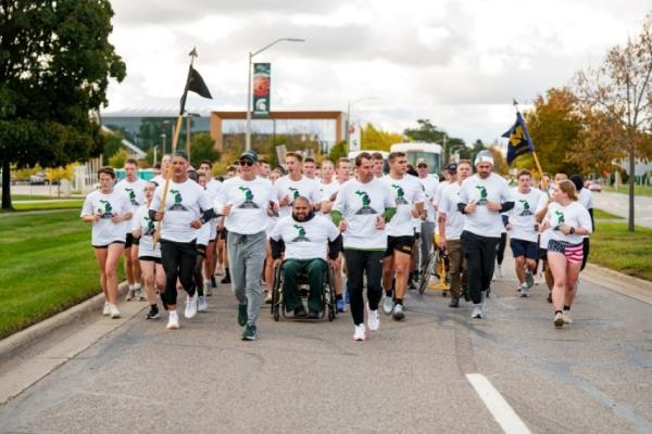 A group of participants traveling on a road, some running and some utilizing mobility devices. Participants are carrying U-M and MSU flags.