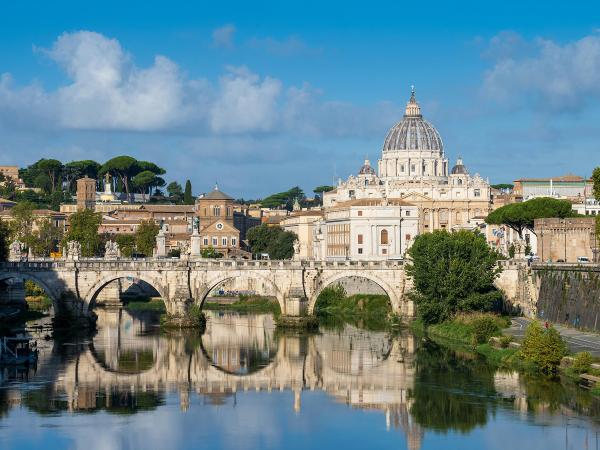View of the Tiber River in Rome, showing a bridge, buildings, and greenery reflected in its surface, under a blue sky.