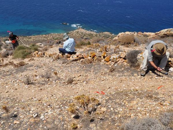 Three archaeologists work at the site of Stelida on a rocky hillside descending toward the bright-blue water of the Aegean Sea.