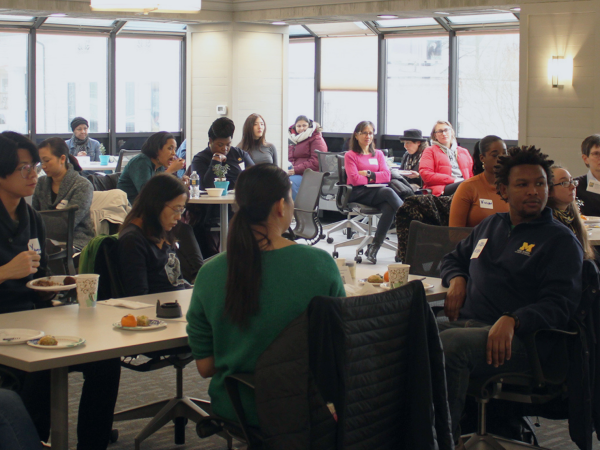 People seated at tables in a bright room, engaged in an event or meeting, with food and drinks on their tables.