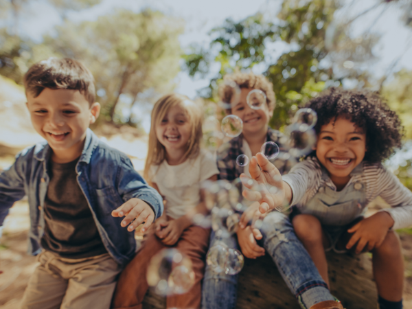 Four children sitting close together outside, laughing and reaching toward bubbles in the air. Sunlight filters through trees in the background.