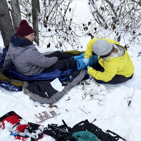 Two participants in a snowy wooded area in winter gear, one participant is laying down on layers of tarp simulating an emergency their leg is being wrapped by another participant in a bright green coat.