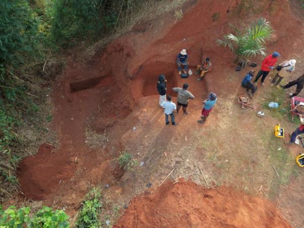 A high-angle drone photograph of an excavation project. Several researchers and field workers stand near cut-out rectangular earth pits and trenches, analyzing soil stratigraphy in a tropical landscape with bright red clay.