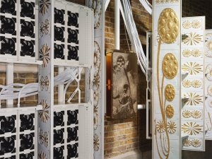 An art installation blending computer server racks, black cooling fans, and white panels decorated with intricate gold floral patterns against a brick wall.