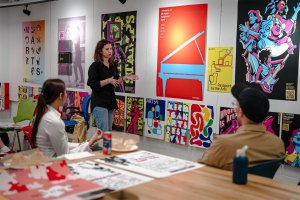 A student discusses their design in front of a large wall containing multiple colorful posters