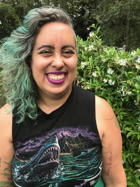 an image of the speaker smiling in front of flowers. they are wearing lipstick and their hair is tinted green.