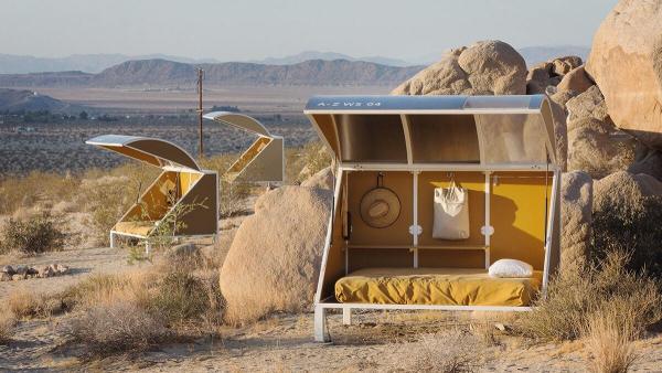 A wide shot of a small architectural shelter with a bed located next to boulders. Mountains are in the distance.