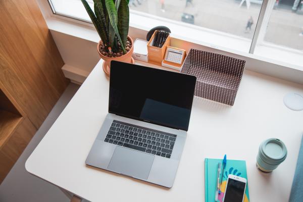 work from home desk setup with a laptop and colorful book, pens, coffee cup, box and plant that overlooks a street/sidewalk with a fair amount of foot traffic