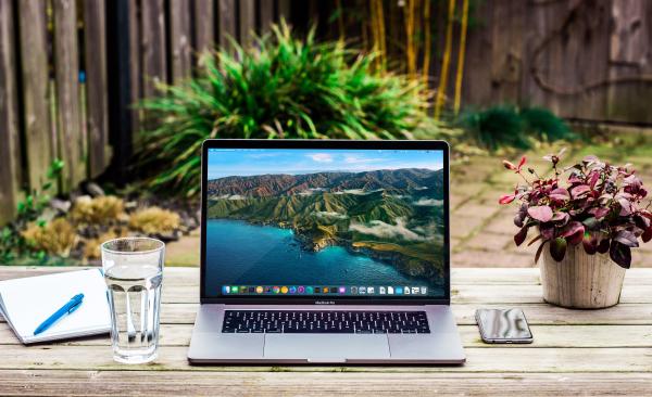 outdoor desk with a plant, laptop, phone, glass of water, pen & paper resting on it (with a bush and a wooden fence in the background)