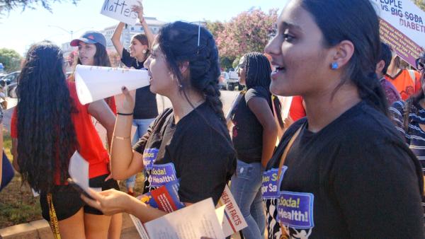 People participating in a health advocacy rally, holding signs and flyers. One person speaks through a megaphone. Trees and buildings are visible in the background.